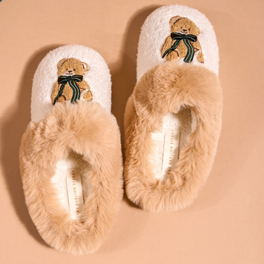 A pair of plush slip-on slippers with beige faux fur trim and embroidered teddy bear faces with dark green bows on the top, shown on a warm beige background. A green 'PRE-PACK' banner is visible in the top-left corner.