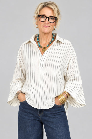 A woman with short blonde hair and glasses wearing a cream and navy blue vertically striped button-down shirt and dark blue jeans, standing inside a boutique shop.
