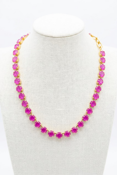 A gold tennis-style necklace with bright pink gemstones displayed on a beige fabric jewelry bust in a retail setting.