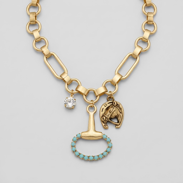 A gold-toned chunky chain necklace with multiple charms, including a small clear rhinestone, a horse-head charm, and a hammer-shaped charm outlined with turquoise stones, displayed on a beige fabric jewelry bust.