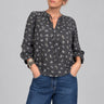 A woman with short gray hair and glasses wearing a dark gray floral print blouse and blue jeans, standing in a boutique store with wooden floors and clothing racks in the background.