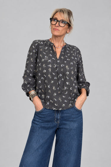 A woman with short gray hair and glasses wearing a dark gray floral print blouse and blue jeans, standing in a boutique store with wooden floors and clothing racks in the background.