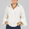 A woman with short blonde hair and glasses wearing a cream and navy blue vertically striped button-down shirt and dark blue jeans, standing inside a boutique shop.
