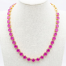 A gold tennis-style necklace with bright pink gemstones displayed on a beige fabric jewelry bust in a retail setting.